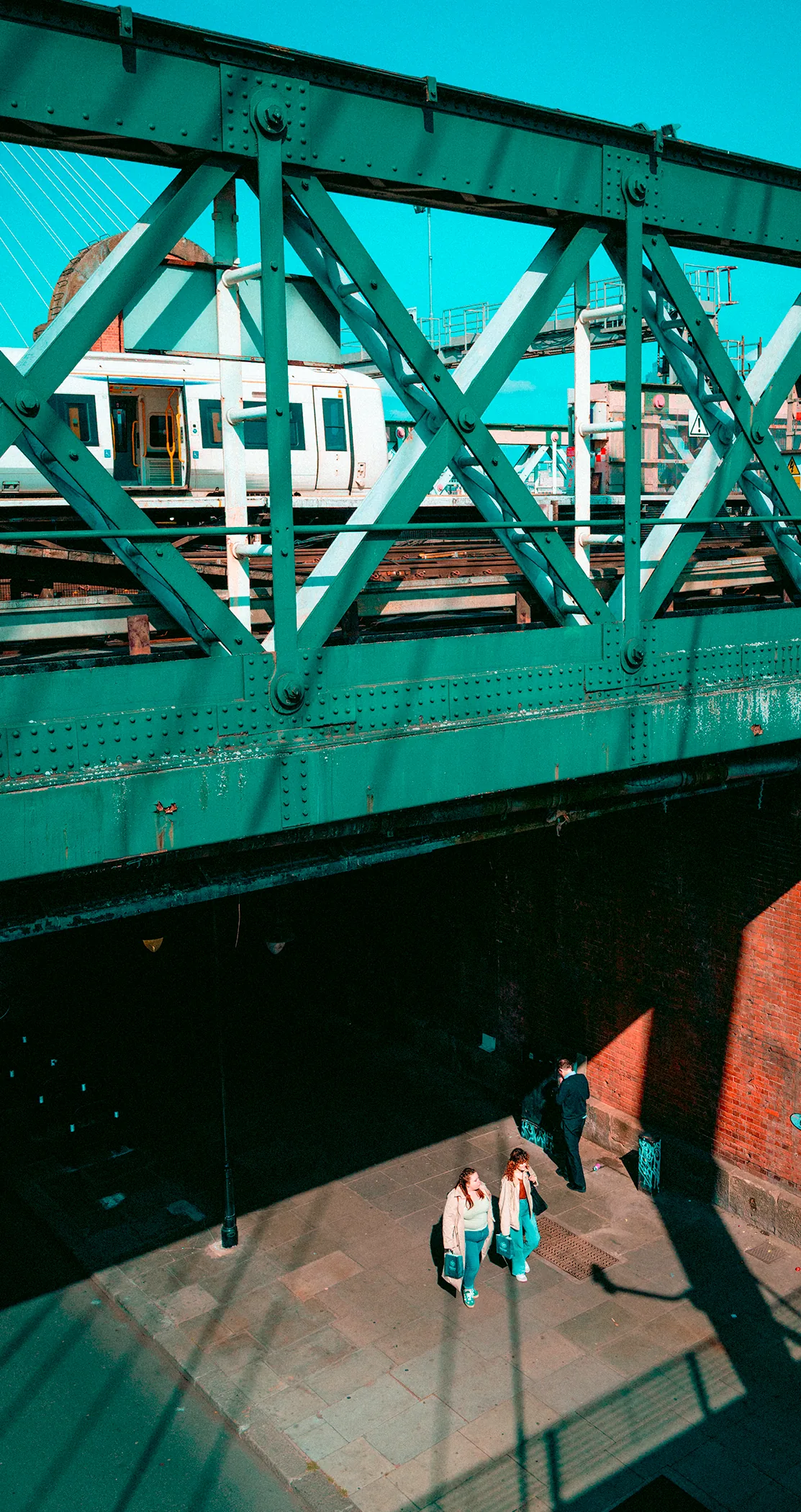 Two people crossing a sunlit square beneath a green steel bridge
