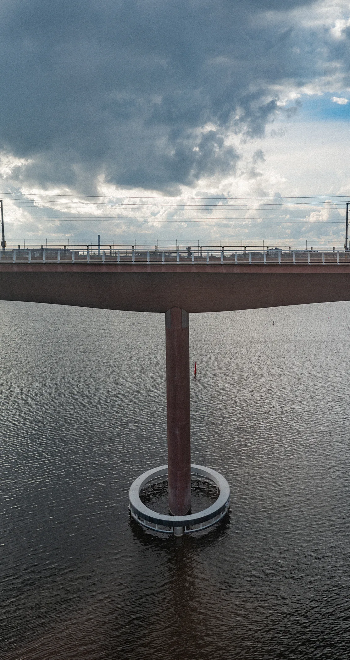 A red road bridge on a single pillar ringed by a fender, over dark water under breaking cloud