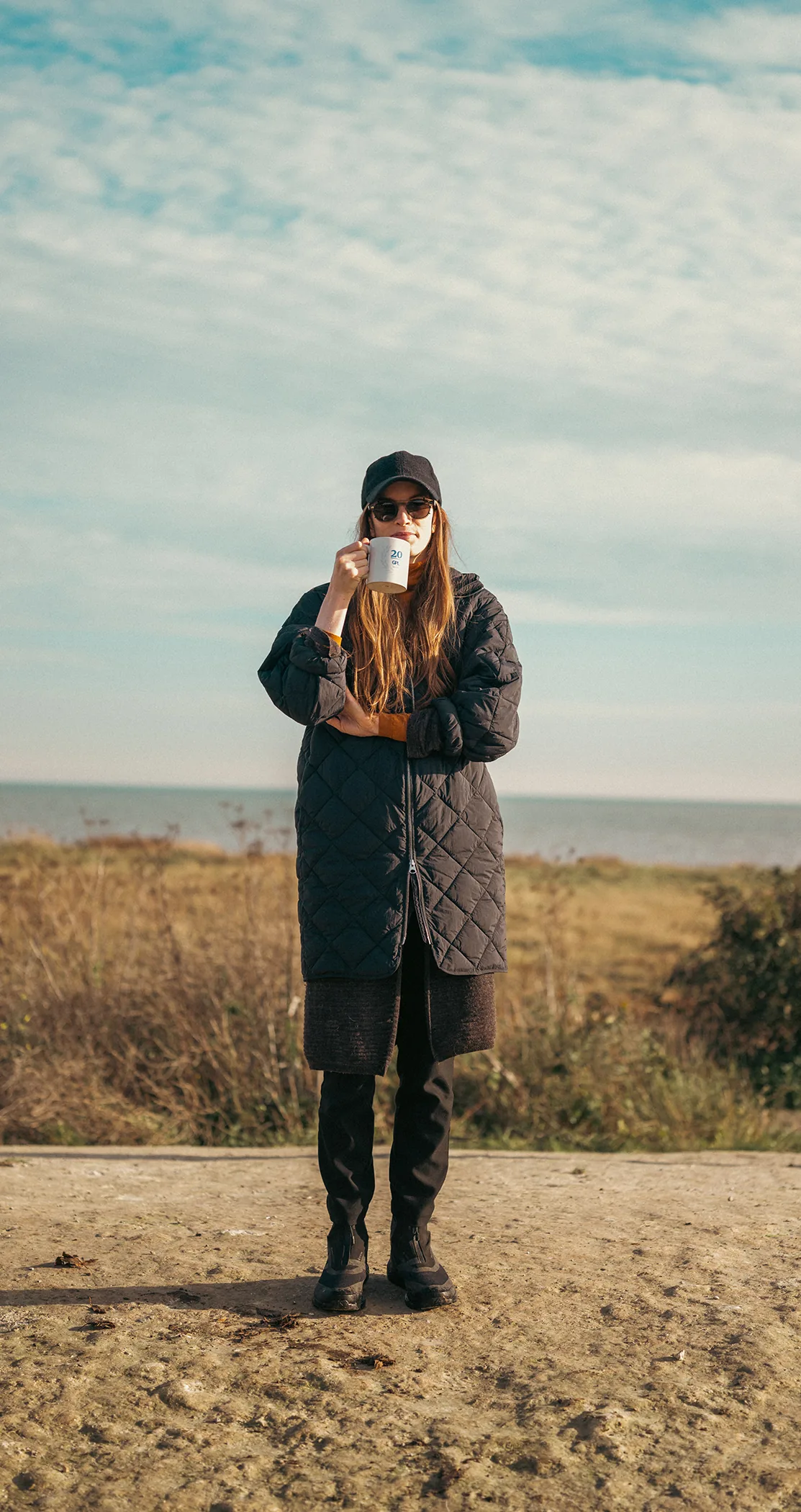 A woman in a long quilted coat sipping from a mug on the dunes, Margate