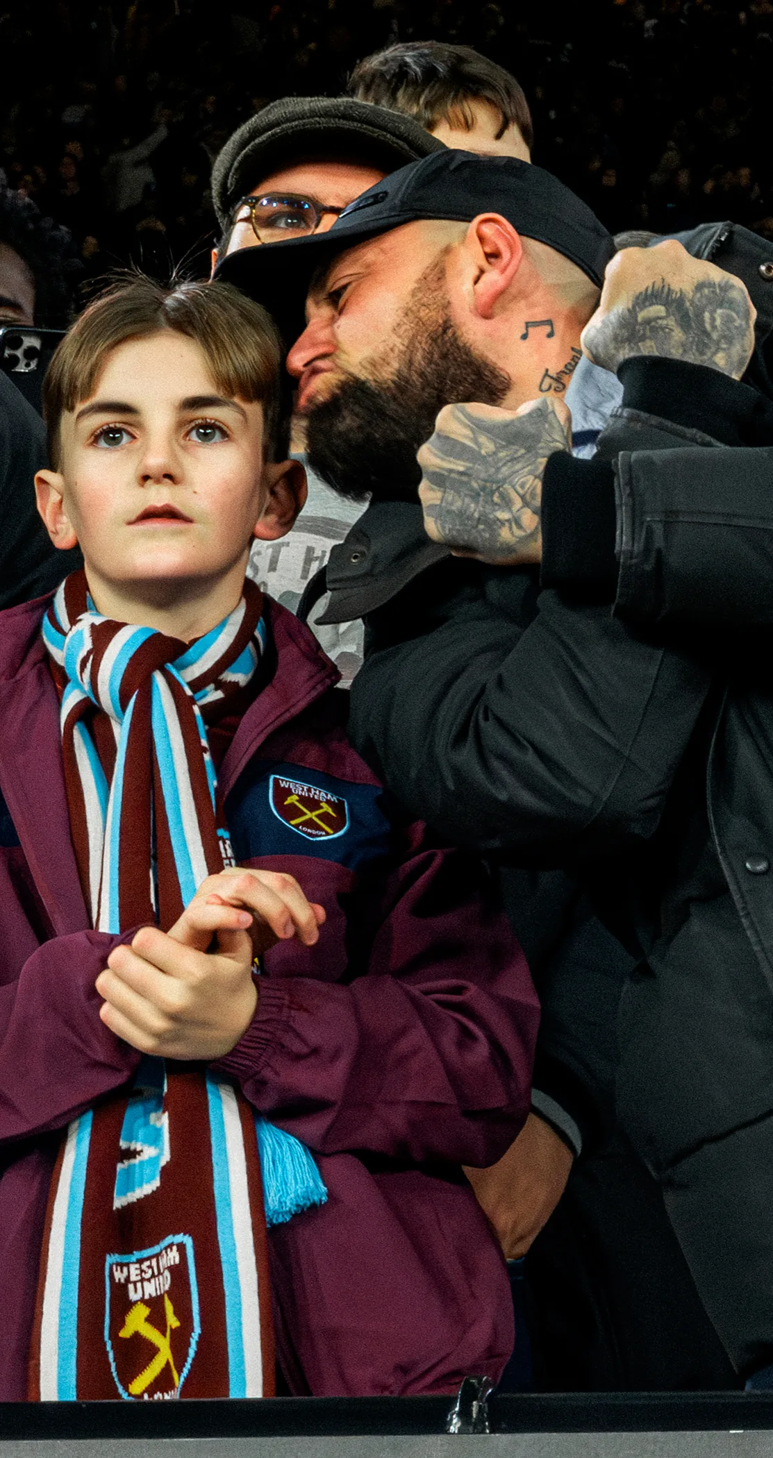 A young West Ham fan in his scarf with a man leaning in behind him